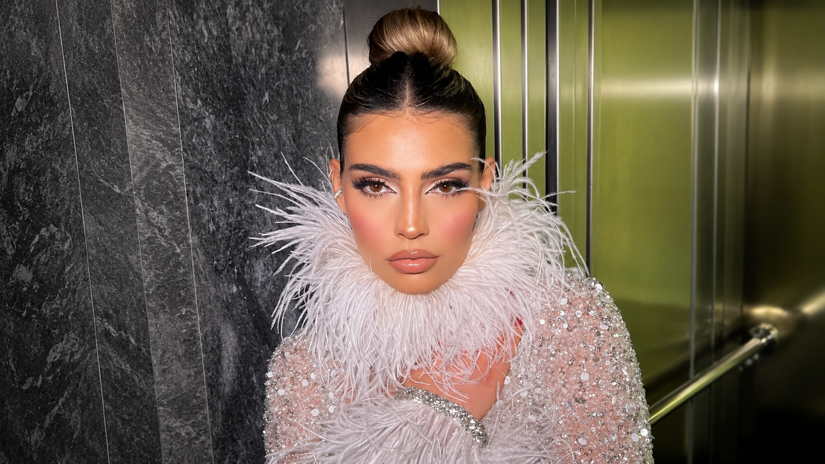 Client with a high sleek bun, bold eye definition and a warm nude lip against a marble and gold backdrop